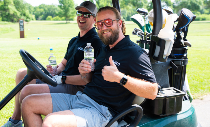Chris Arnold and Joel Larson riding in a golf cart with their thumbs up.