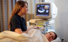 A healthcare professional does an echocardiogram on a person lying on an exam table under a blanket while heart images appear on a screen.